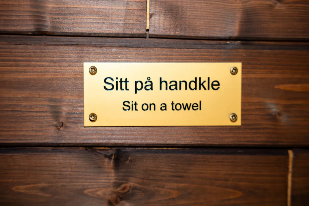 Bilete av messingskilt med tekst som lyder "Sitt på handkle" og på engelsk: "Sit on a towel". Trebakgrunn.