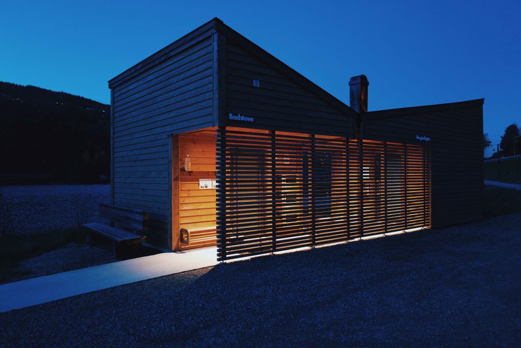 Bilete av badstova tatt på kveldstid i mørkret. Lys skin ut mellom spilene i vindfanget.